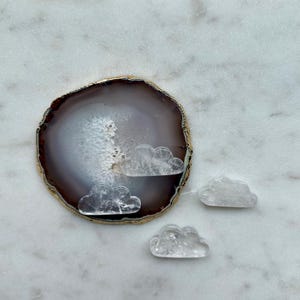 May include: A natural agate slice with a gold-painted edge, featuring three small, clear quartz cloud carvings. The agate is set on a white marble surface with a scalloped edge. The clouds are intricately carved.