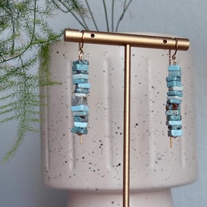 May include: A pair of dangle earrings featuring stacked, rectangular, light blue and mottled grey-blue stone beads on thin gold-toned metal wires. The beads vary in shade and texture, creating a vertical, column-like appearance for each earring.