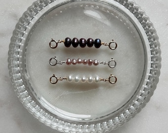 May include: Three jewelry extenders featuring small, round pearls in dark purple, light pink, and white. Each extender has a circular spring ring clasp at both ends, connected by thin metal wire. The top extender is gold-toned, the middle is silver-toned, and the bottom is gold-toned.
