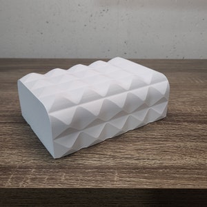 May include: A rectangular white object with a repeating geometric pattern of raised triangular pyramids across its top and front surfaces. The object has a smooth, matte finish and a solid, block-like structure with rounded edges along the top.