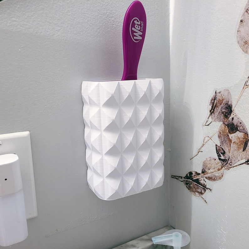 May include: A white, geometric-patterned holder with a purple Wet Brush hairbrush. The holder is mounted on a light gray wall. The brush is inserted into the holder, with the handle visible.
