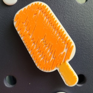 May include: Orange popsicle-shaped wall decor made of small, interlocking pieces. The popsicle is white with an orange design and a yellow stick.