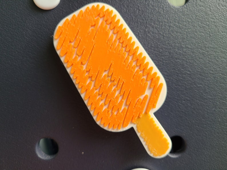 May include: Orange popsicle-shaped wall decor made of small, interlocking pieces. The popsicle is white with an orange design and a yellow stick.