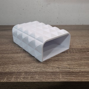 May include: A white rectangular holder with a hollow interior and a textured exterior surface featuring a repeating pattern of raised triangular pyramids. The object has a smooth finish and a wide, open rectangular opening on one end.