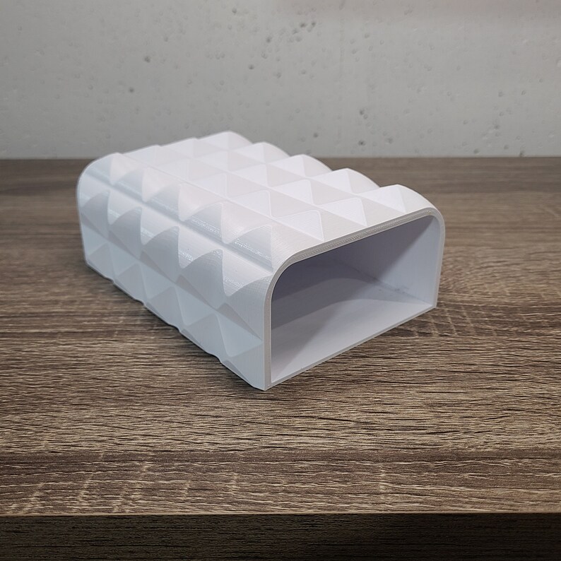May include: A white rectangular holder with a hollow interior and a textured exterior surface featuring a repeating pattern of raised triangular pyramids. The object has a smooth finish and a wide, open rectangular opening on one end.