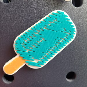 May include: A teal blue popsicle with a textured surface and an orange stick. The popsicle is on a dark gray background.