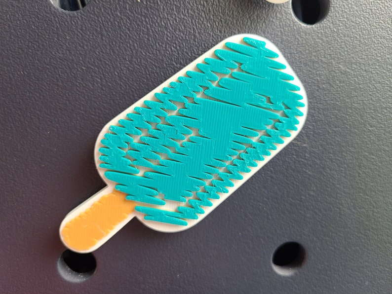 May include: A teal blue popsicle with a textured surface and an orange stick. The popsicle is on a dark gray background.