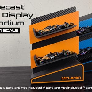 May include: Diecast F1 display podium in black, orange, and blue, designed for 1:64 scale model cars. Features the McLaren and Alpine logos. The podium has multiple tiers to display the model cars.