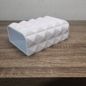 May include: A white, rectangular soap dish with a geometric, textured design. The dish has a curved front and a series of diamond-shaped patterns across its surface. The interior is smooth and open, designed to hold a bar of soap.