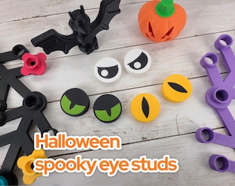 Może przedstawiać: Zestaw elementów dekoracyjnych o tematyce Halloween, w tym czarny nietoperz, pomarańczowa dynia i różne kolczyki z przerażającymi oczami w kolorze białym, zielonym i żółtym. Widoczny jest również tekst "Halloween spooky eye studs".