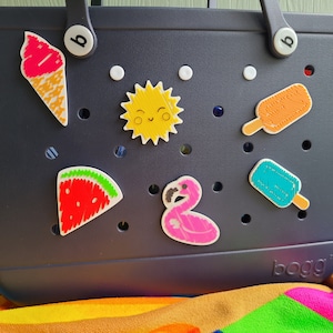 May include: A dark blue tote bag with a white polka dot pattern and various colorful embroidered patches, including a watermelon, ice cream cone, flamingo, sun, and popsicles. The bag has the word "bogg" printed on the bottom.
