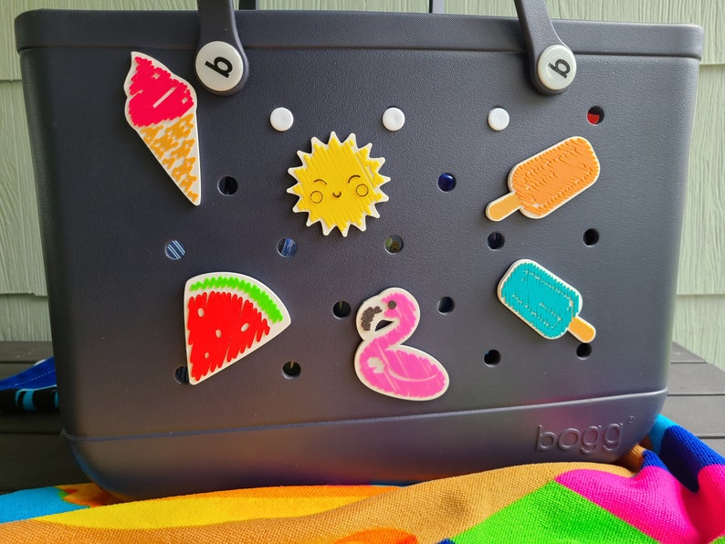 May include: A dark blue tote bag with a white polka dot pattern and various colorful embroidered patches, including a watermelon, ice cream cone, flamingo, sun, and popsicles. The bag has the word "bogg" printed on the bottom.