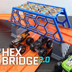 May include: A blue and orange Hot Wheels track set with a hexagonal bridge. The track is orange and the bridge is blue with orange hexagonal cutouts. Three toy monster trucks are on the track. The text "HEX BRIDGE 2.0" is printed on the track.