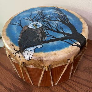 May include: A round wooden drum with a rawhide head featuring a hand-painted scene of a bald eagle perched on a dark tree branch against a blue sky. The drum body is constructed from polished wood and secured with light-colored leather lacing.