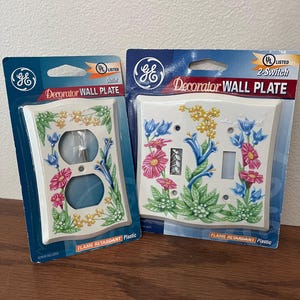 May include: Two GE Decorator Wall Plates. One is an outlet cover, the other a 2-switch cover. Both are white with floral designs in pink, blue, and yellow. The packaging reads "FLAME RETARDANT Plastic".