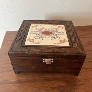 May include: A small, rectangular wooden jewelry box with a decorative tile top. The tile features a cream background with an oval brown center and red and brown swirling patterns. The box has a dark brown finish and a metal clasp.