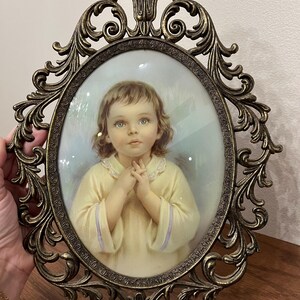 May include: An oval-shaped framed portrait of a child with light brown hair and blue eyes, wearing a yellow dress. The frame is ornate, with a bronze finish and decorative scrollwork. The child's hands are clasped together in front of their chest.