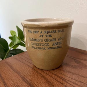 Peut inclure: Un pot en grès beige avec un bord épais et un corps conique. L'avant présente un texte imprimé en bleu foncé qui indique : YOU GET A SQUARE DEAL AT THE FARMERS GRAIN AND LIVESTOCK ASS'N. COLERIDGE, NEBRASKA. La surface a une finition émaillée lisse.
