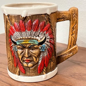 May include: A vintage ceramic mug featuring a Native American chief design. The mug has a brown, textured exterior with a detailed face and headdress in red, white, and blue. The handle is also brown and textured, and the rim is white.