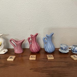 May include: A collection of miniature ceramic figurines, including a white swan with gold accents, pink and blue pitchers, and two teacups with saucers. The items are arranged on a wooden surface.