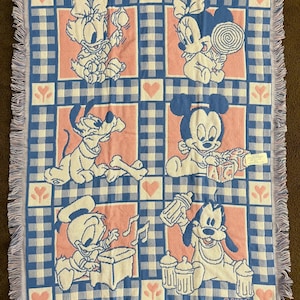 May include: A baby blanket featuring Disney characters Daisy Duck, Minnie Mouse, Pluto, Mickey Mouse, Donald Duck, and Goofy. The blanket has a blue and white checkered pattern with pink squares and fringe. The Disney characters are in a baby-like style.