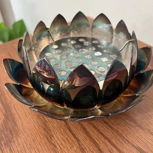 May include: A metallic lotus flower-shaped bowl with a dark, iridescent finish and a removable circular insert featuring a grid of small, evenly spaced holes. The petals are layered and pointed, showing a gradient of dark blue, green, and bronze tones.
