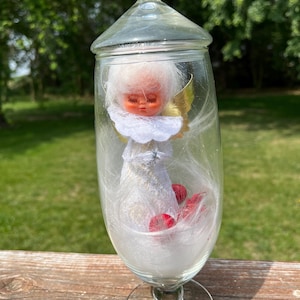 May include: A vintage glass cloche with a clear, rounded base and a lid, housing a small angel figurine. The angel has a white dress, gold wings, and a serene expression. Red heart-shaped accents are visible inside the glass.