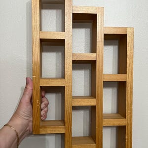 May include: A light brown wooden wall shelf featuring a staggered, multi-level design with nine rectangular compartments. The structure is composed of three vertical sections of varying heights, each containing three open storage cubbies.
