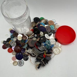 May include: A clear plastic jar with a red lid, spilling out a large collection of colorful buttons of various sizes and shapes.