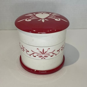 May include: White ceramic jar with a red lid. The lid has a white heart and floral design. The jar has a red band around the top and bottom and a pink floral design around the middle.