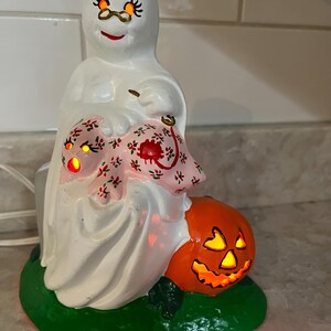 May include: A white ceramic Halloween ghost figure wearing glasses and holding a pink floral item. An orange jack-o'-lantern sits at the base. The decoration is on a green base and has an internal light.