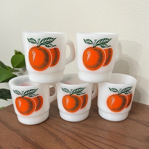 May include: A set of five white milk glass mugs, each featuring a vibrant orange fruit graphic with dark green leaves. The mugs have a stackable design with a rounded handle on the side, showcasing a consistent citrus print on the front surface.