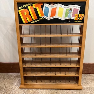 May include: A vintage wooden retail display rack for Rit Tints and Dyes featuring five shelves with thin metal dividers. The top sign displays the text All Purpose Rit Tints and Dyes, The Finest Dye That Money Can Buy, and 25 cents Big Box.