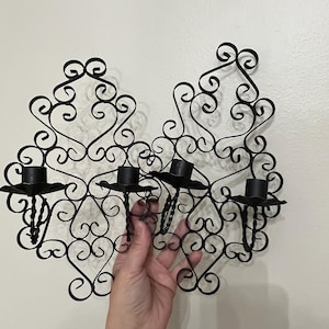 May include: A black metal wall sconce featuring two symmetrical, ornate scrollwork panels. The design includes four cylindrical candle holders, each supported by a twisted metal stem and a flat, petal-shaped base, arranged horizontally across the frame.