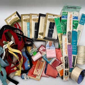 May include: An assortment of sewing supplies including zips, ribbon, trim, and fabric. The supplies are packaged in various colours and patterns. The text on the packaging reads "Slip Straps", "Metal Trouser Zipper", "Shapeflex Non-Fusible Interfacing", and "Dritz Lace Trim".