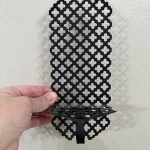 May include: Black metal wall sconce with a decorative quatrefoil pattern. The sconce has a small shelf and a bracket for mounting. The design is symmetrical and the metal has a matte finish.