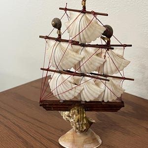 May include: A decorative seashell ship with white shell sails, brown wooden masts, and red rigging. The ship features three snail shell accents and sits on a base made of seashells. A unique nautical-themed home decor item.