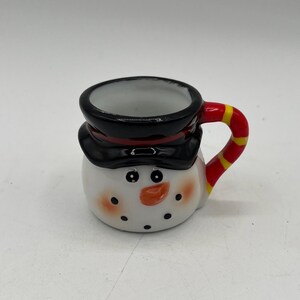 May include: A ceramic snowman-shaped mug with a black top hat and a red and yellow striped handle. The mug features a white body, black dot eyes, a carrot nose, and rosy cheeks. A festive holiday item.