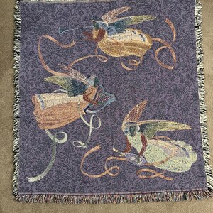 Puede incluir: Una manta tejida con un fondo morado y un patr&oacute;n repetitivo. La manta presenta tres figuras de &aacute;ngeles con alas y cintas que fluyen en tonos beige, azul y dorado. Los bordes est&aacute;n acabados con un borde con flecos.