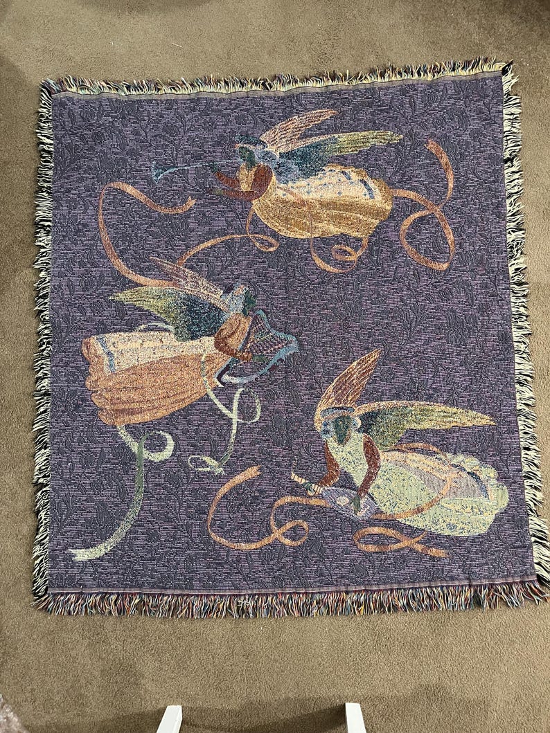 Puede incluir: Una manta tejida con un fondo morado y un patr&oacute;n repetitivo. La manta presenta tres figuras de &aacute;ngeles con alas y cintas que fluyen en tonos beige, azul y dorado. Los bordes est&aacute;n acabados con un borde con flecos.