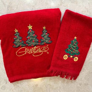 May include: Two red Christmas towels. One towel features three embroidered Christmas trees with gold stars and the word "Greetings". The other towel has a single Christmas tree design with a gold star.