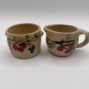 May include: A set of two small ceramic creamer and sugar bowls with a floral design. The bowls are beige with green, red, and blue painted vines and flowers.