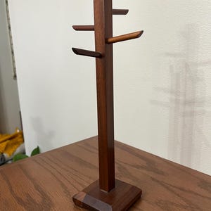 May include: A dark brown wooden mug tree with a square base and four horizontal arms. The tree is made of solid wood and has a simple, modern design. It is designed to hold mugs or cups.