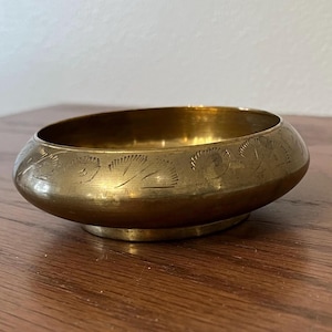 May include: A small, antique brass bowl with a rounded shape and a slightly raised base. The bowl's exterior features an etched floral pattern around the rim. The brass has a warm, golden hue, suggesting age and use. This decorative bowl is suitable for small items.