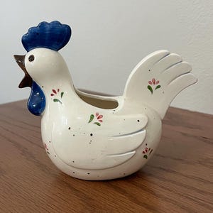 May include: A ceramic rooster-shaped planter with a white body, blue comb, and brown beak. The planter has a small opening at the top and is decorated with small red and green floral accents and black speckles. The rooster is sitting on a wooden surface.
