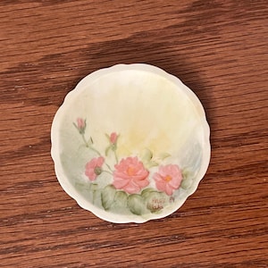 May include: A small, round, off-white dish with a scalloped edge. The dish features a hand-painted floral design with pink roses, green leaves, and buds. The artist's signature and the year '89 are visible. The background is a soft yellow.