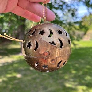 May include: A vintage brass sphere ornament with star and crescent moon cutouts. The sphere is suspended by a delicate gold chain. The ornament has a hinged opening and a warm, aged patina, suggesting it is an antique.