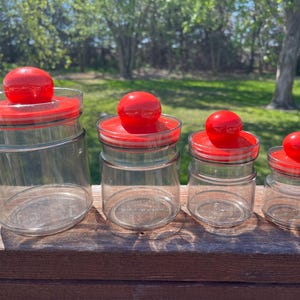 May include: Four glass canisters with red lids. The canisters are clear glass and the lids are a bright red colour. The canisters are arranged in a row from largest to smallest.