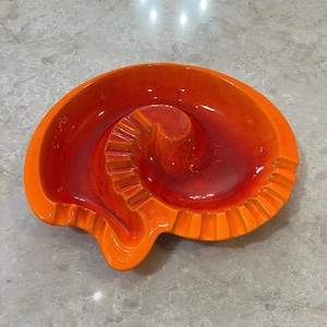 May include: A circular ceramic ashtray featuring a vibrant orange and deep red gradient glaze. The design includes a central raised spiral pattern with integrated grooves along the outer rim, creating a distinct shell-like appearance.