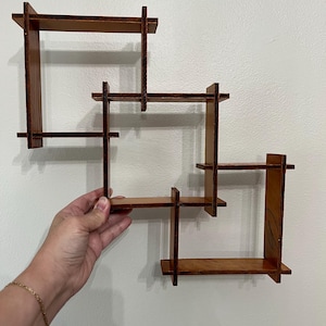 May include: A wall-mounted wooden shelving unit featuring three interconnected square frames with a dark brown finish. The structure consists of multiple horizontal and vertical planks joined together to create several open display compartments.
